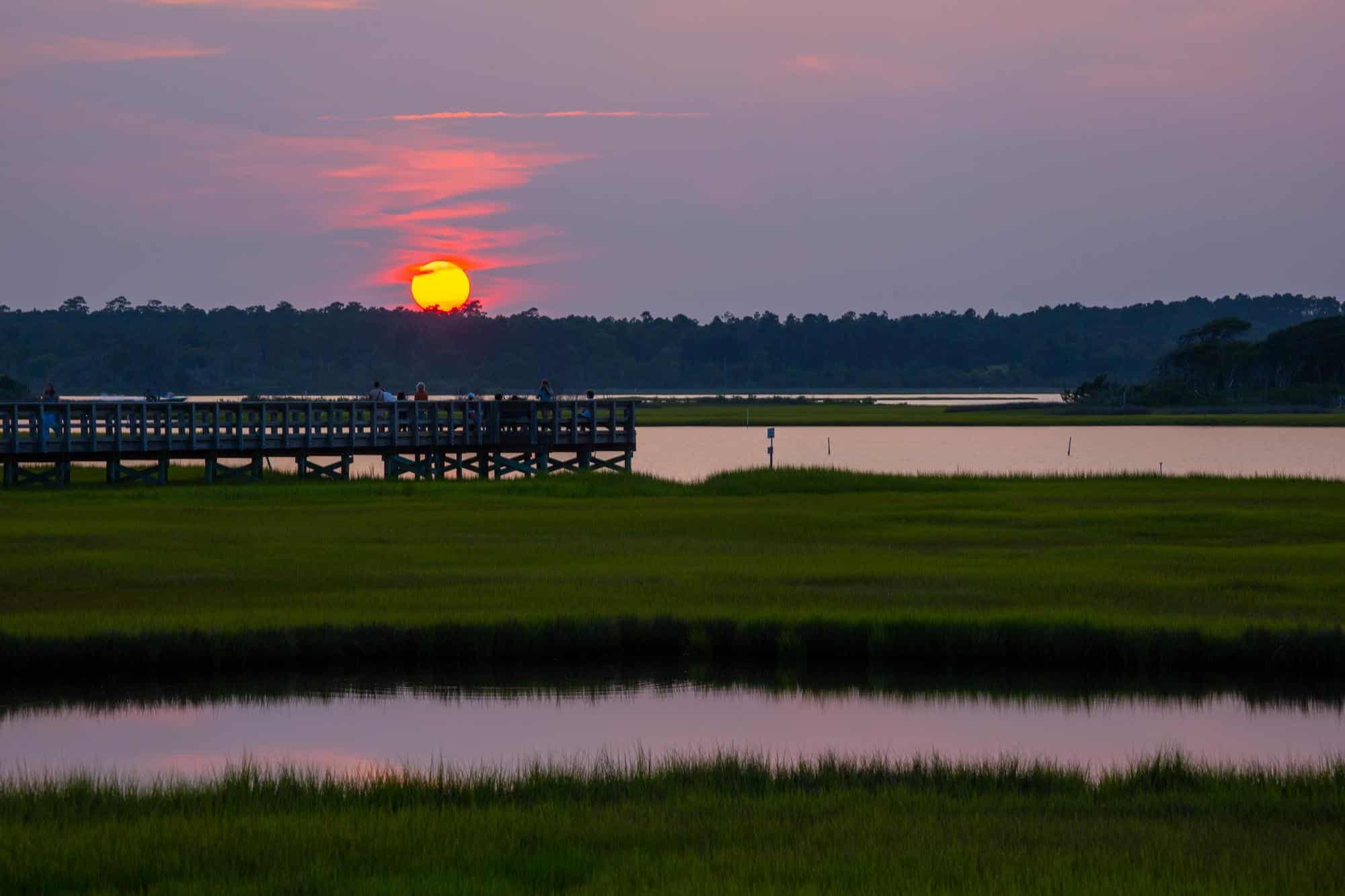 A body of water with a dock, grass and trees in the background during sunset.